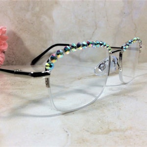 May include: Silver-toned eyeglasses featuring a semi-rimless frame design. The upper portion of the frames is embellished with a row of iridescent, multi-colored crystals that catch the light. The temples are thin and metallic with black tips.
