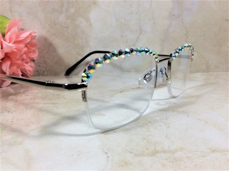 May include: Silver-toned eyeglasses featuring a semi-rimless frame design. The upper portion of the frames is embellished with a row of iridescent, multi-colored crystals that catch the light. The temples are thin and metallic with black tips.