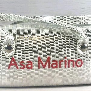 May include: Silver handbag with a textured pattern and a top handle. The name "Asa Marino" is printed in red on the front of the bag. The bag has silver hardware.
