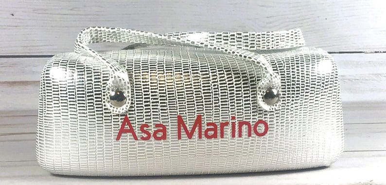 May include: Silver handbag with a textured pattern and a top handle. The name "Asa Marino" is printed in red on the front of the bag. The bag has silver hardware.