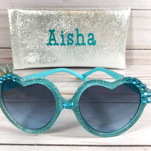 May include: Teal heart-shaped sunglasses with rhinestones and a gold crown detail. The sunglasses come with a silver glitter case with the name "Aisha" embroidered on it.
