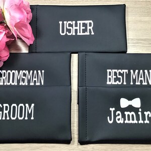 May include: Four black fabric pouches with white text. The pouches are labeled "Usher", "Groomsman", "Groom", and "Best Man" with a bow tie and the name "Jamir".