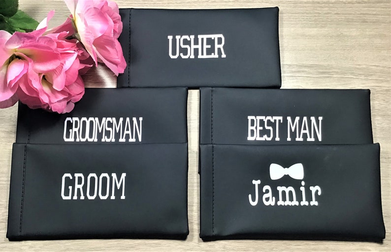 May include: Four black fabric pouches with white text. The pouches are labeled "Usher", "Groomsman", "Groom", and "Best Man" with a bow tie and the name "Jamir".