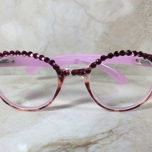 K&ouml;nnte beinhalten: Rosa Brille mit dekorativem Design. Die Fassungen sind hellrosa mit einem Schildpattmuster an den unteren R&auml;ndern. Die Oberseite der Fassungen ist mit Reihen kleiner, dunkelrosa Strasssteine verziert.