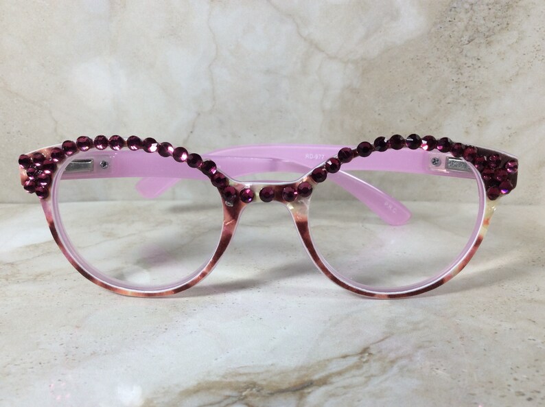 K&ouml;nnte beinhalten: Rosa Brille mit dekorativem Design. Die Fassungen sind hellrosa mit einem Schildpattmuster an den unteren R&auml;ndern. Die Oberseite der Fassungen ist mit Reihen kleiner, dunkelrosa Strasssteine verziert.