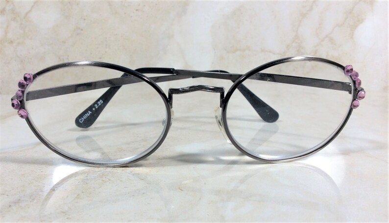Puede incluir: Gafas ovaladas con montura de metal gris oscuro. Las patillas est&aacute;n adornadas con peque&ntilde;os brillantes rosas. Las lentes son transparentes y el texto "CHINA +2.25" est&aacute; impreso en la patilla.