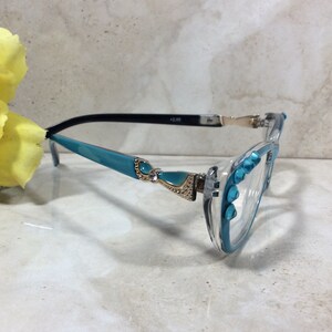 May include: Clear-framed reading glasses with turquoise accents and decorative embellishments. The frames feature turquoise detailing on the arms and around the lenses, with small, sparkling accents. The glasses have a +2.00 magnification.