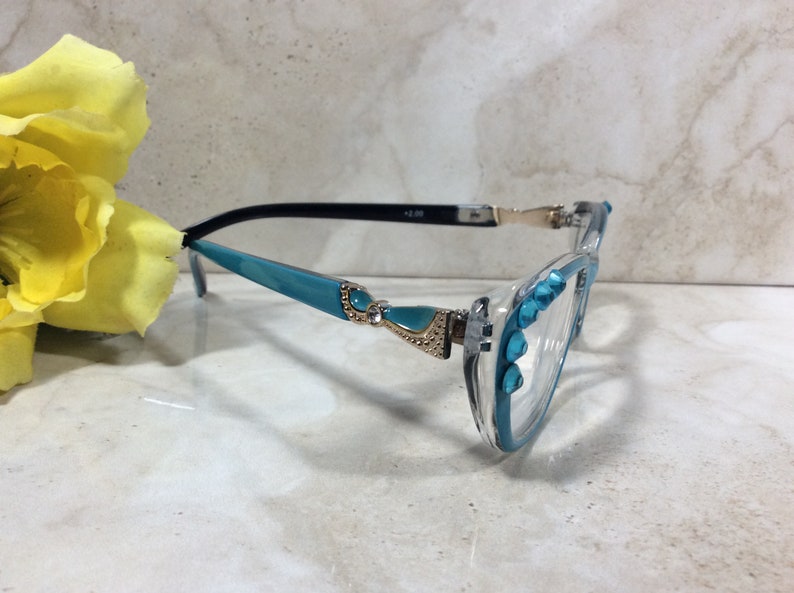 May include: Clear-framed reading glasses with turquoise accents and decorative embellishments. The frames feature turquoise detailing on the arms and around the lenses, with small, sparkling accents. The glasses have a +2.00 magnification.