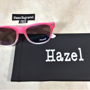 May include: Pink and white ombre sunglasses with a black case. The case has the name "Hazel" printed on it in white.