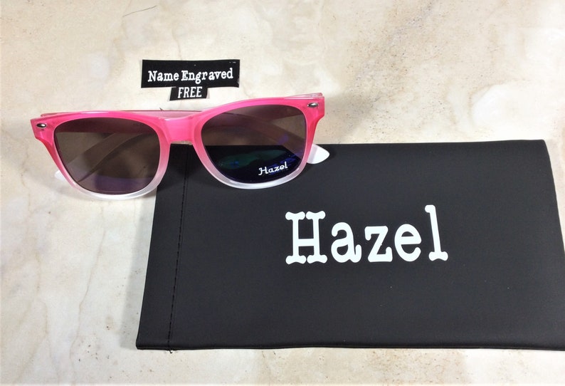 May include: Pink and white ombre sunglasses with a black case. The case has the name "Hazel" printed on it in white.