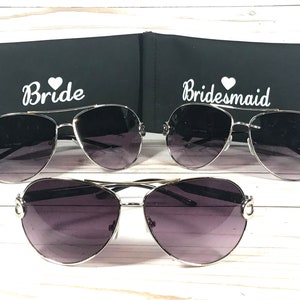 May include: Three pairs of aviator sunglasses with gradient lenses and silver frames. Two pairs are displayed with black cases, one labeled "Bride" and the other "Bridesmaid" in white script. The third pair is displayed below.