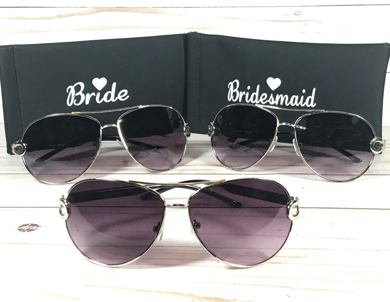 May include: Three pairs of aviator sunglasses with gradient lenses and silver frames. Two pairs are displayed with black cases, one labeled "Bride" and the other "Bridesmaid" in white script. The third pair is displayed below.