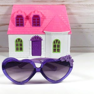 May include: Purple heart-shaped sunglasses with a decorative flower detail on the side. The sunglasses have a dark lens and are made of plastic.