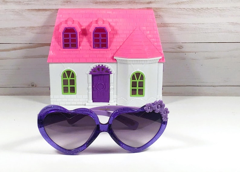 May include: Purple heart-shaped sunglasses with a decorative flower detail on the side. The sunglasses have a dark lens and are made of plastic.