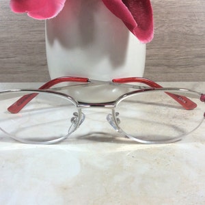 Pu&ograve; includere: Occhiali con montatura argentata, aste rosse e gemme rosa decorative sugli angoli esterni. Le lenti sono trasparenti e gli occhiali hanno i naselli. Il testo "China" &egrave; visibile sull'asta.