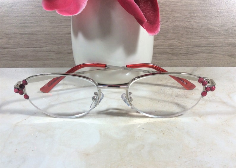 Pu&ograve; includere: Occhiali con montatura argentata, aste rosse e gemme rosa decorative sugli angoli esterni. Le lenti sono trasparenti e gli occhiali hanno i naselli. Il testo "China" &egrave; visibile sull'asta.