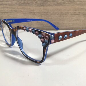 May include: Eyeglasses with a blue and tortoiseshell frame, decorated with pink and blue rhinestones. The temples are brown with blue rhinestones. The glasses have a rectangular shape and are for vision correction.