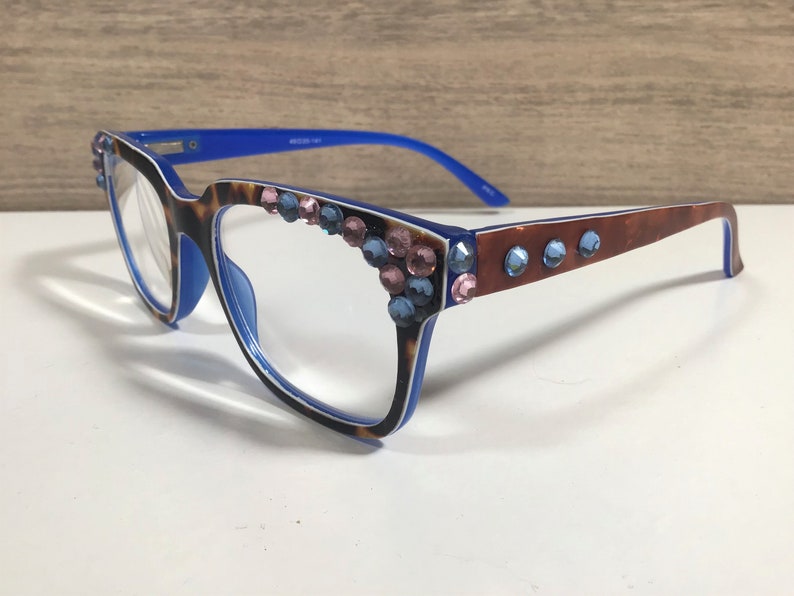 May include: Eyeglasses with a blue and tortoiseshell frame, decorated with pink and blue rhinestones. The temples are brown with blue rhinestones. The glasses have a rectangular shape and are for vision correction.