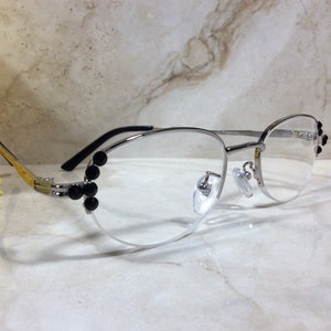May include: Silver-framed eyeglasses with clear lenses. The frames feature black embellishments on the sides and near the nose bridge. The temples are black, and the hinges are gold. A yellow flower is in the background.