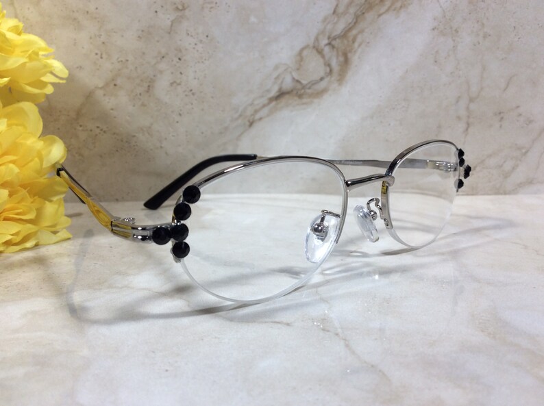 May include: Silver-framed eyeglasses with clear lenses. The frames feature black embellishments on the sides and near the nose bridge. The temples are black, and the hinges are gold. A yellow flower is in the background.