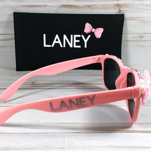 Op de afbeelding: Roze zonnebril met strass en een roze strik aan de zijkant. De zonnebril heeft de naam "LANEY" op de pootjes.