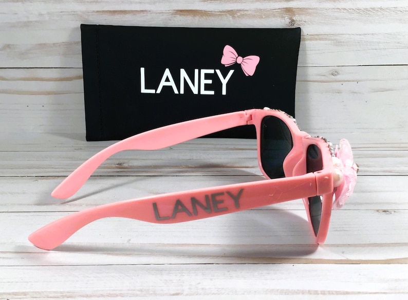 Op de afbeelding: Roze zonnebril met strass en een roze strik aan de zijkant. De zonnebril heeft de naam "LANEY" op de pootjes.