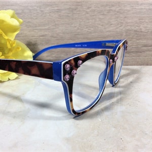 May include: Blue and tortoise shell eyeglasses with pink rhinestone accents. The frames have a blue and white trim. The lenses are clear. A yellow flower is in the background.