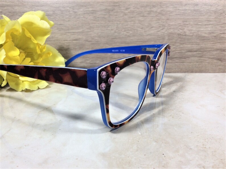 May include: Blue and tortoise shell eyeglasses with pink rhinestone accents. The frames have a blue and white trim. The lenses are clear. A yellow flower is in the background.