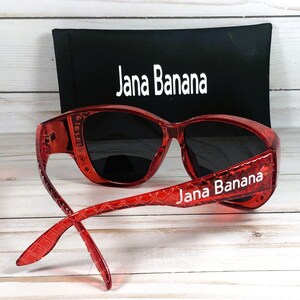 May include: Red sunglasses with black lenses and the text "Jana Banana" on the temples. A black case with the text "Jana Banana" is behind the sunglasses. The sunglasses are on a light wood surface.