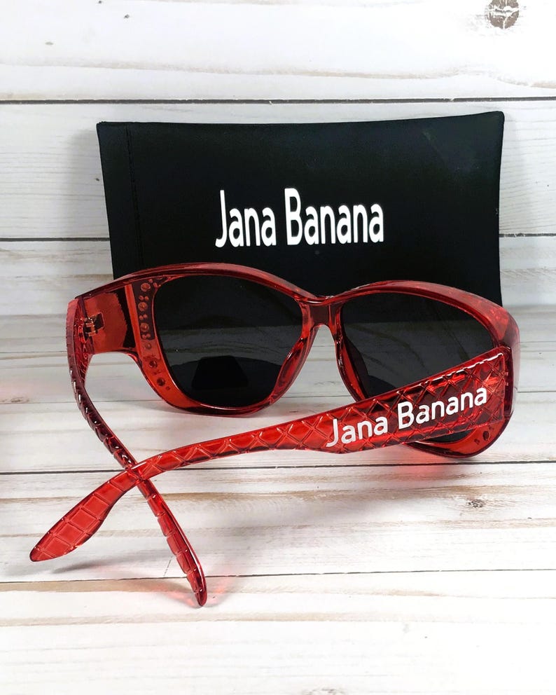 May include: Red sunglasses with black lenses and the text "Jana Banana" on the temples. A black case with the text "Jana Banana" is behind the sunglasses. The sunglasses are on a light wood surface.
