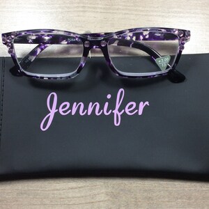 K&ouml;nnte beinhalten: Eine Lesebrille mit lila und wei&szlig;em Muster und schwarzem Rahmen liegt auf einem schwarzen Etui. Auf dem Etui steht der Name "Jennifer" in hellvioletter Schrift. Die Brille hat eine St&auml;rke von +3,00.