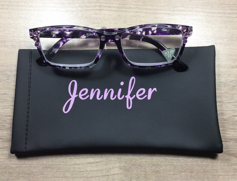 K&ouml;nnte beinhalten: Eine Lesebrille mit lila und wei&szlig;em Muster und schwarzem Rahmen liegt auf einem schwarzen Etui. Auf dem Etui steht der Name "Jennifer" in hellvioletter Schrift. Die Brille hat eine St&auml;rke von +3,00.