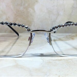 May include: Silver-framed eyeglasses with a half-rim design. The top of the frame is adorned with a row of small, sparkling rhinestones. The lenses are clear, and the nose pads are visible. The glasses are set against a soft, blurred background.