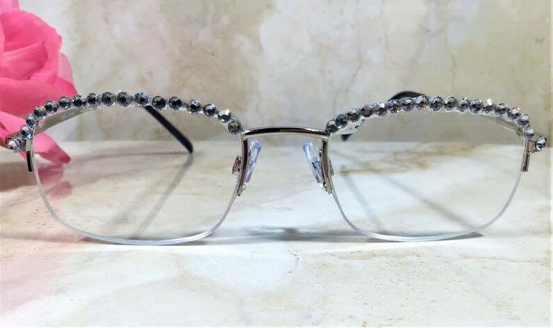 May include: Silver-framed eyeglasses with a half-rim design. The top of the frame is adorned with a row of small, sparkling rhinestones. The lenses are clear, and the nose pads are visible. The glasses are set against a soft, blurred background.