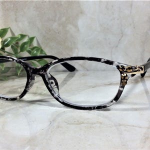 Peut inclure: Lunettes de vue ovales avec une monture au motif mouchet&eacute; noir et blanc. Les branches sont reli&eacute;es &agrave; la face par des charni&egrave;res en m&eacute;tal dor&eacute; orn&eacute;es de petits cristaux transparents &agrave; facettes dispos&eacute;s en grappe florale de chaque c&ocirc;t&eacute;.