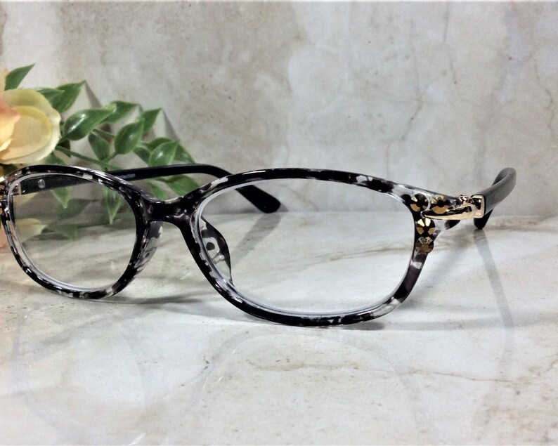 Peut inclure: Lunettes de vue ovales avec une monture au motif mouchet&eacute; noir et blanc. Les branches sont reli&eacute;es &agrave; la face par des charni&egrave;res en m&eacute;tal dor&eacute; orn&eacute;es de petits cristaux transparents &agrave; facettes dispos&eacute;s en grappe florale de chaque c&ocirc;t&eacute;.