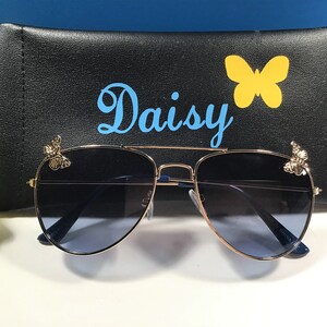 Op de afbeelding: Gouden aviator zonnebril met blauwe glazen en vlinderaccenten op de pootjes. De zonnebril zit in een zwarte etui met het woord "Daisy" erop gedrukt.