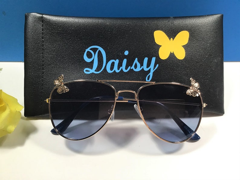 Op de afbeelding: Gouden aviator zonnebril met blauwe glazen en vlinderaccenten op de pootjes. De zonnebril zit in een zwarte etui met het woord "Daisy" erop gedrukt.