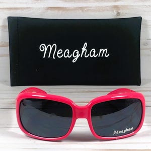 May include: Hot pink sunglasses with dark lenses and the name "Meagham" printed on the right lens. A black case with the name "Meagham" in white script is behind the sunglasses.