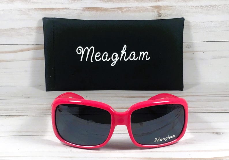 May include: Hot pink sunglasses with dark lenses and the name "Meagham" printed on the right lens. A black case with the name "Meagham" in white script is behind the sunglasses.