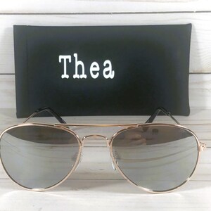 May include: Aviator sunglasses with a thin gold-tone metal frame and reflective silver lenses. Behind the sunglasses sits a rectangular black protective case featuring the white text Thea printed in a clean, sans-serif font on the front.
