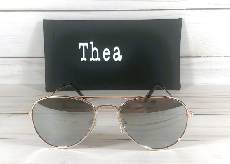 May include: Aviator sunglasses with a thin gold-tone metal frame and reflective silver lenses. Behind the sunglasses sits a rectangular black protective case featuring the white text Thea printed in a clean, sans-serif font on the front.