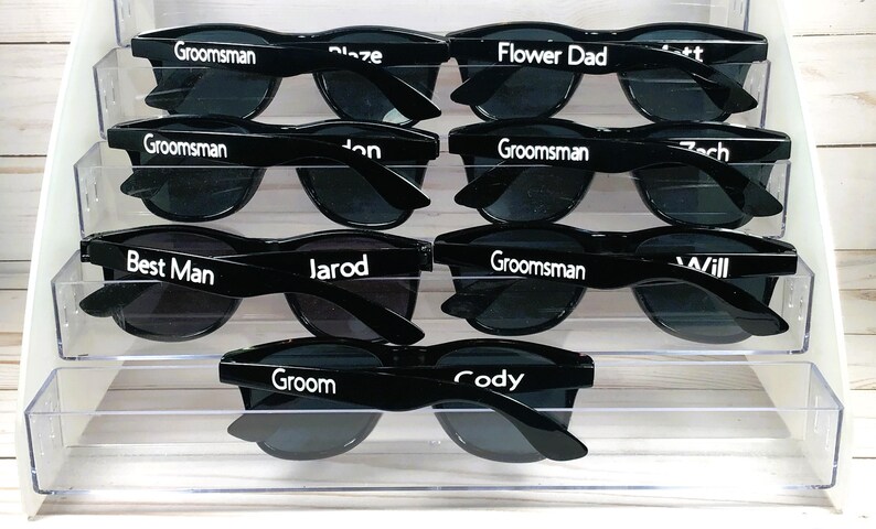 May include: A set of eight black sunglasses with white text on the lenses. The text on the sunglasses reads "Groomsman", "Best Man", "Groom", "Flower Dad", "Cody", "Zach", "Jarod", and "Will".