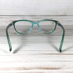 May include: A pair of eyeglasses featuring a translucent teal frame with a gradient effect that transitions into a darker green at the temples. The lenses are clear and oval-shaped, connected by a thin bridge, with curved arms extending outward.