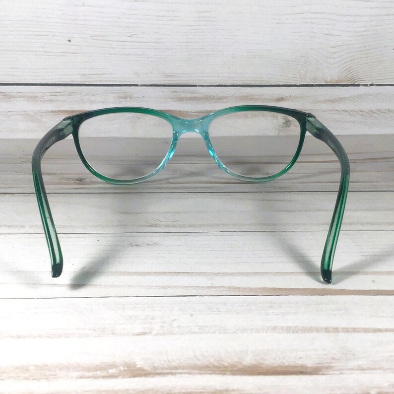 May include: A pair of eyeglasses featuring a translucent teal frame with a gradient effect that transitions into a darker green at the temples. The lenses are clear and oval-shaped, connected by a thin bridge, with curved arms extending outward.