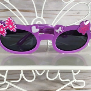 May include: Purple sunglasses with dark lenses, decorated with white and pink floral and heart-shaped accents. The sunglasses are displayed on a white, decorative stand, perfect for children's accessories.