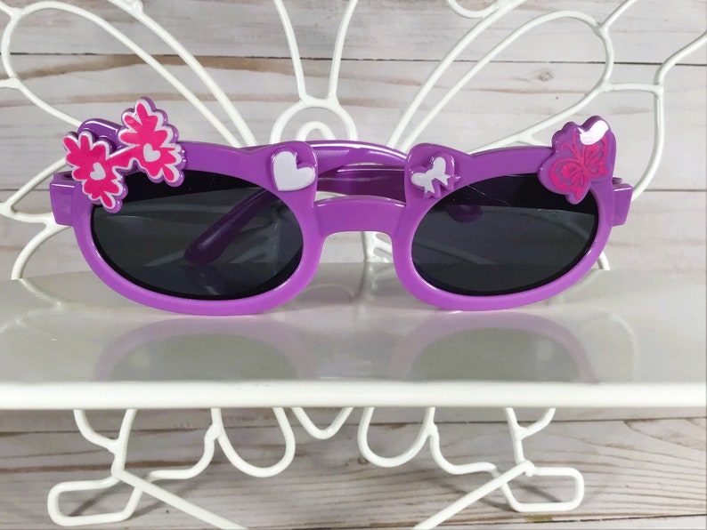 May include: Purple sunglasses with dark lenses, decorated with white and pink floral and heart-shaped accents. The sunglasses are displayed on a white, decorative stand, perfect for children's accessories.