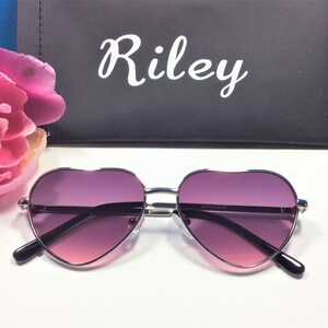 May include: Silver heart-shaped sunglasses with purple gradient lenses. The sunglasses are on a white surface with a pink flower and a black case with the name "Riley" printed on it.