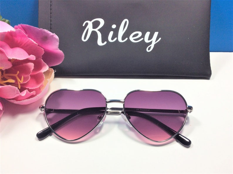 May include: Silver heart-shaped sunglasses with purple gradient lenses. The sunglasses are on a white surface with a pink flower and a black case with the name "Riley" printed on it.