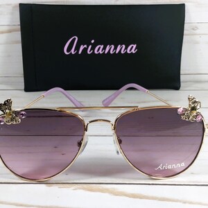 Op de afbeelding: Gouden aviator zonnebril met paarse glazen en vlinderversieringen op de pootjes. De zonnebril heeft de naam "Arianna" op het rechter glas.